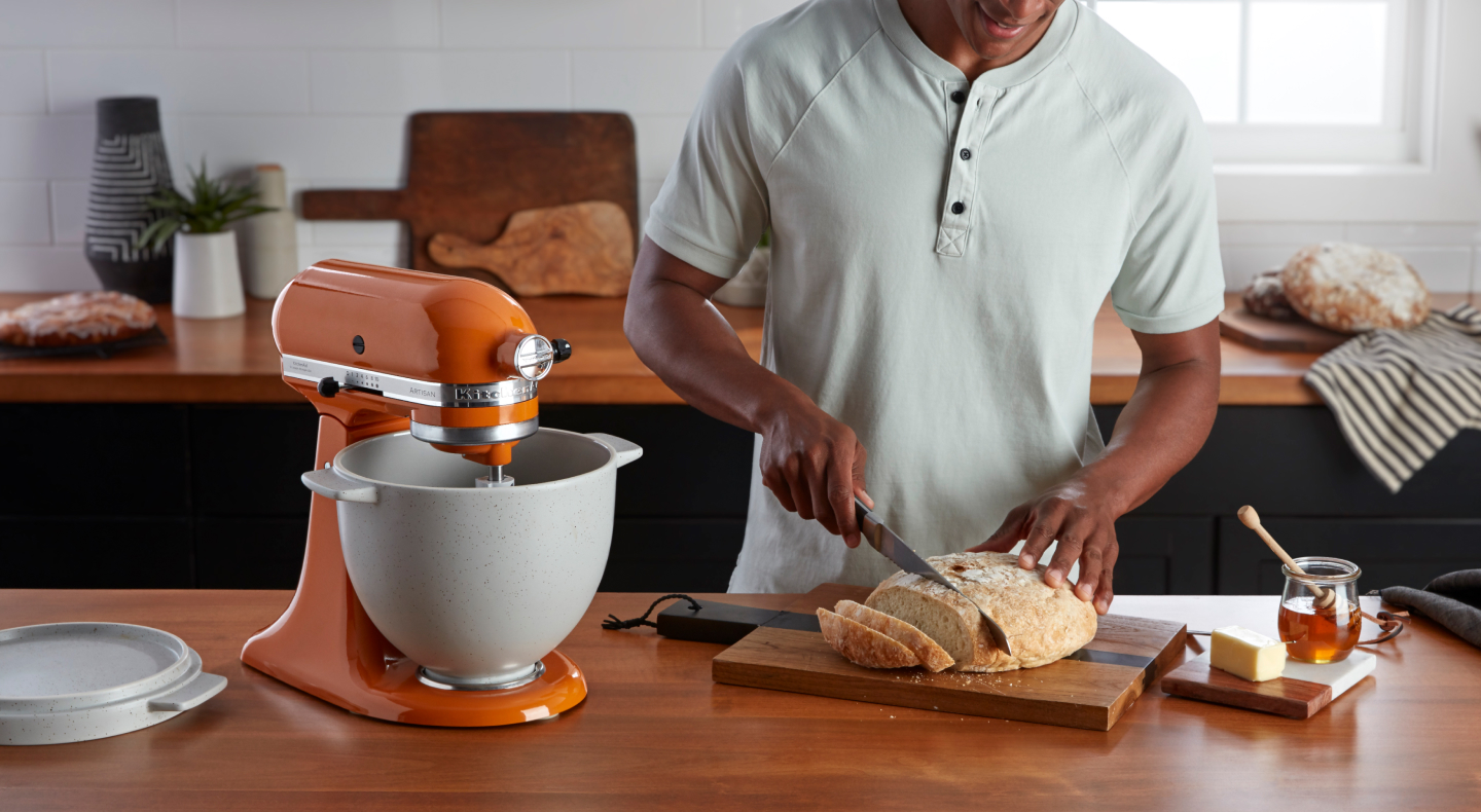 A person slicing homemade bread next to an orange KitchenAid®  Stand Mixer on the counter of a modern kitchen