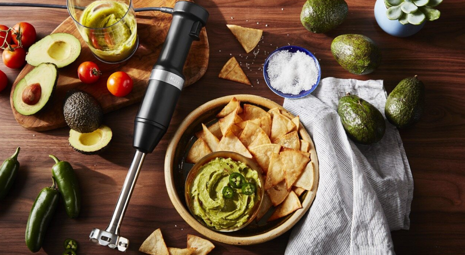 Immersion blender laying beside bowl of guacamole and chips surrounded by guacamole ingredients  Immersion blender laying beside bowl of guacamole and chips surrounded by guacamole ingredients