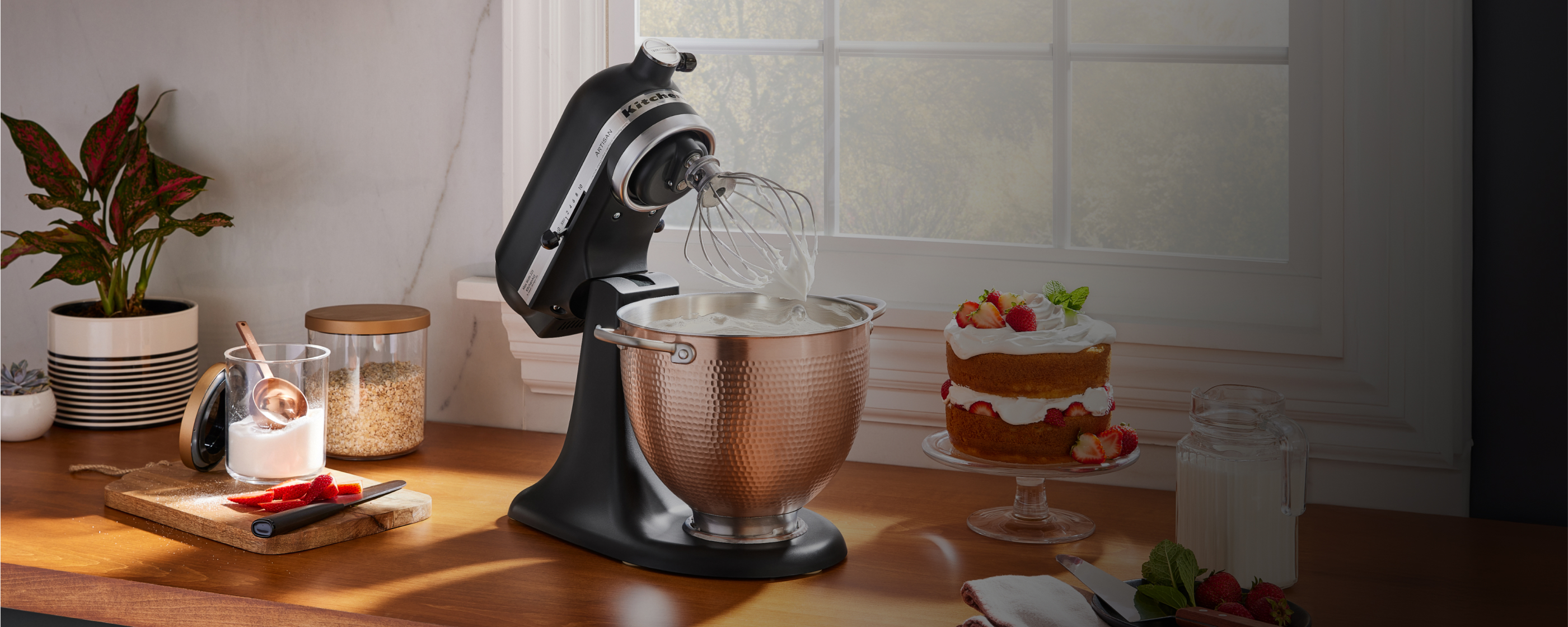 A black KitchenAid® stand mixer with a copper bowl on the counter, near a baked cake and other baking ingredients. 