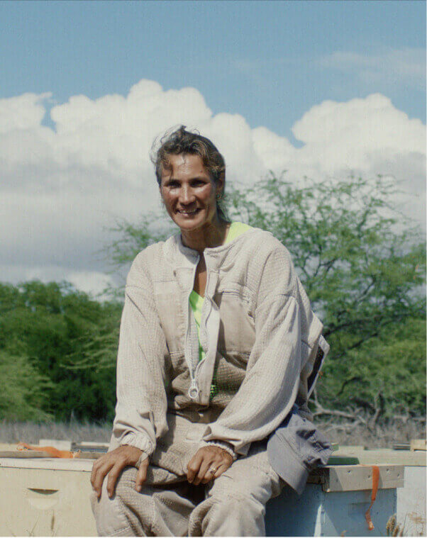 A woman in a beekeeping suit sitting down, smiling for the camera.