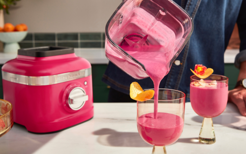 A person pouring a blended hibiscus drink into stylish cups with orange peel garnishes.