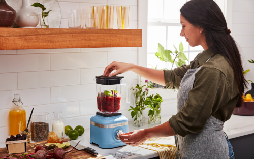 A woman blending fresh vegetables in a KitchenAid® blender in a modern kitchen.