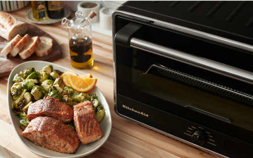 Air fryer salmon in a serving dish with Brussel sprouts next to a KitchenAid® countertop oven with air fry.