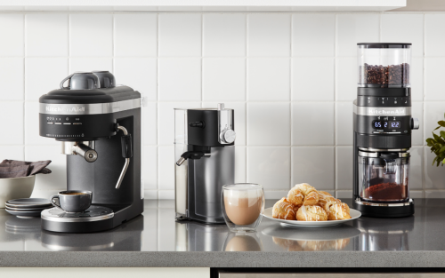 A KitchenAid® espresso machine and burr coffee grinder in a modern kitchen next to a frothy cappuccino and pastries.