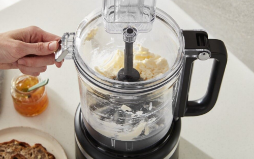 A person opening the lid of a black food processor with butter