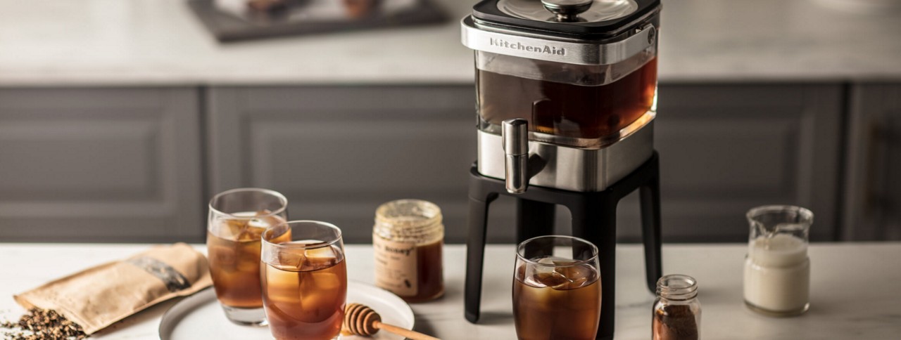 Cold brew coffee setup on kitchen counter including KitchenAid® cold brew coffee maker, three cups of cold brew, milk, honey and cinnamon.