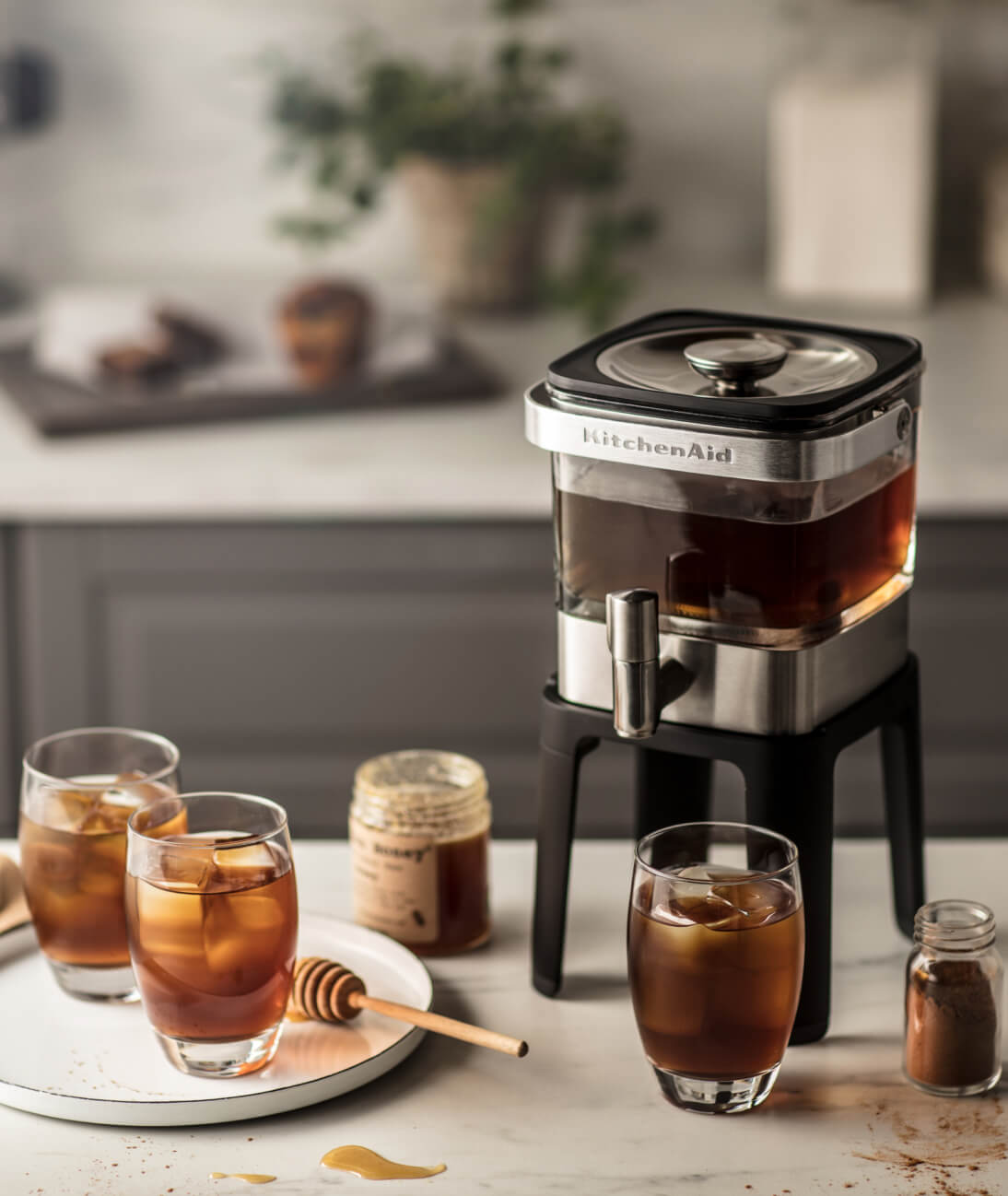 Cold brew coffee setup on kitchen counter including KitchenAid® cold brew coffee maker, three cups of cold brew, milk, honey and cinnamon.