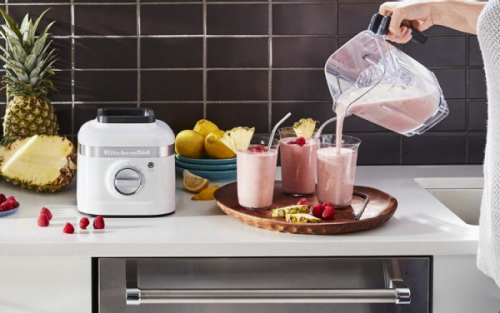 White countertop with KitchenAid® stand mixer and KitchenAid® blender surrounded by fruits - person is pouring smoothies into three glasses