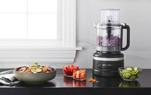 Black food processor on counter surrounded by processed vegetables and a bowl of salad