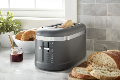 A matte black toaster on a counter with bagels, bread and jam