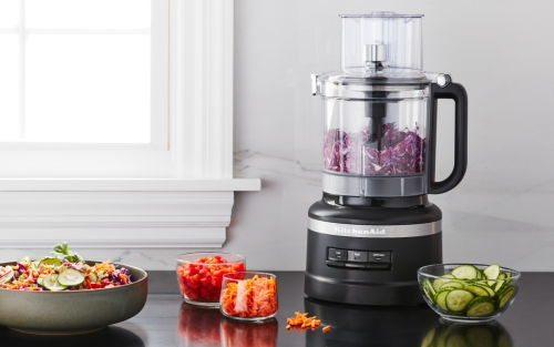 Black KitchenAid® food processor full of shredded red onion next to prepared veggies on the countertop