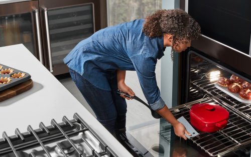 A woman pulling a Dutch oven out of a wall oven surrounded by a variety of baked goods