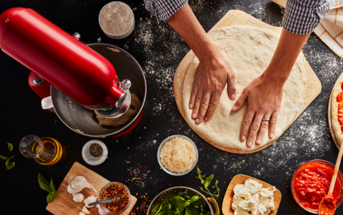 Maker flattening out pizza dough on countertop with KitchenAid® stand mixer and pizza ingredients