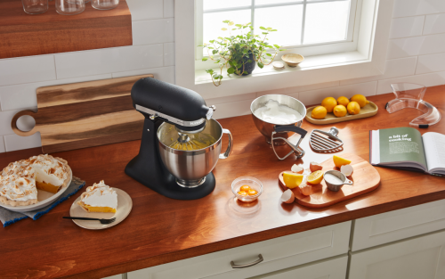 A KitchenAid® stand mixer next to a lemon meringue pie and fresh lemon slices on a cutting board in a modern kitchen. 