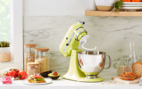 A KitchenAid® stand mixer with a dough hook accessory on a modern kitchen counter next to fresh vegetables for making a sandwich.