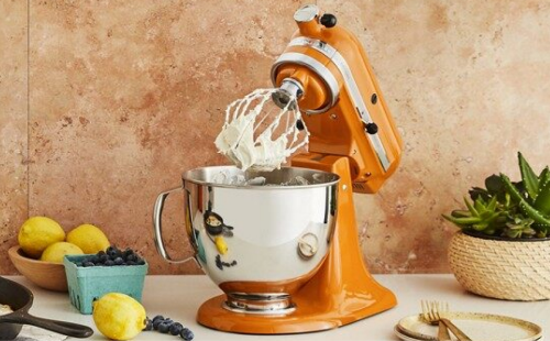 An orange KitchenAid® stand mixer, fruit and a cast iron pan.