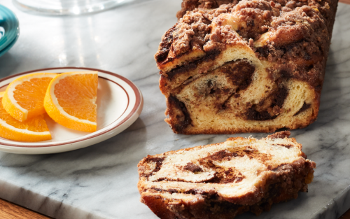 A partially sliced loaf of cinnamon babka on a marble cutting board next to a plate of orange slices