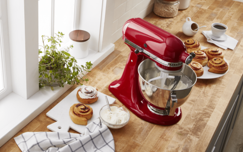 Red KitchenAid® Stand Mixer on a counter surrounded by cinnamon rolls in the process of being iced individually