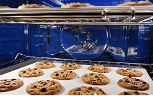 The blue interior of a wall oven, with two racks of chocolate chip cookies baking.