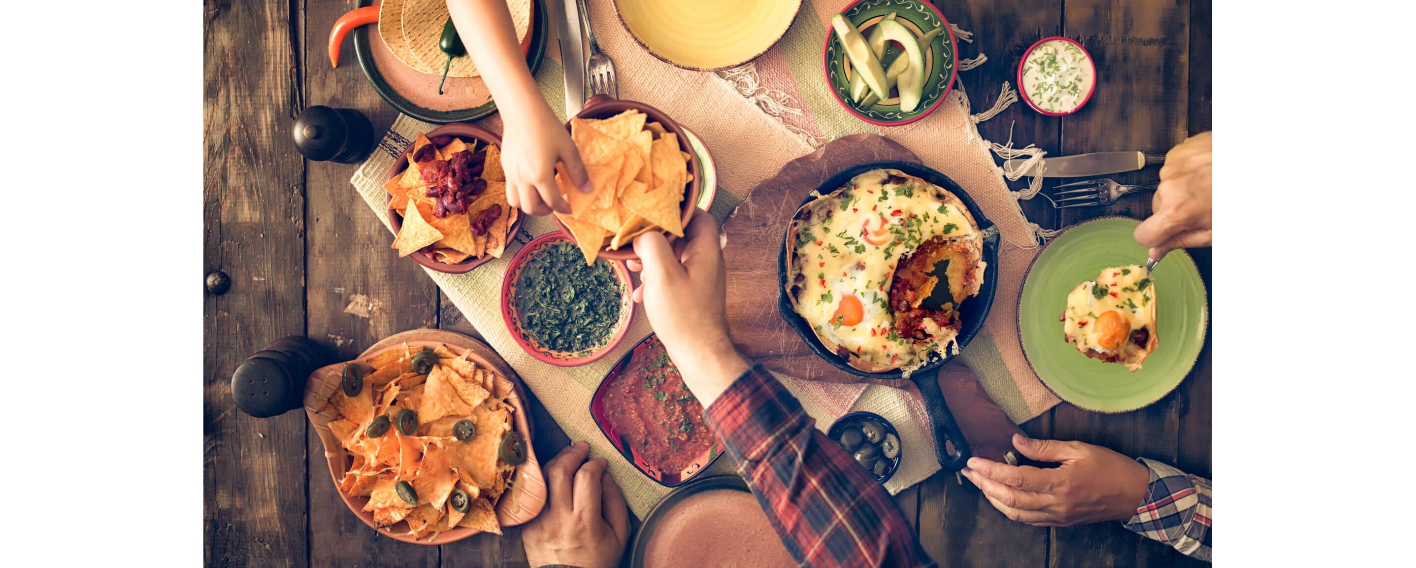 People gathered over a wooden table set with bowls of tortilla chips, dips and salsas.