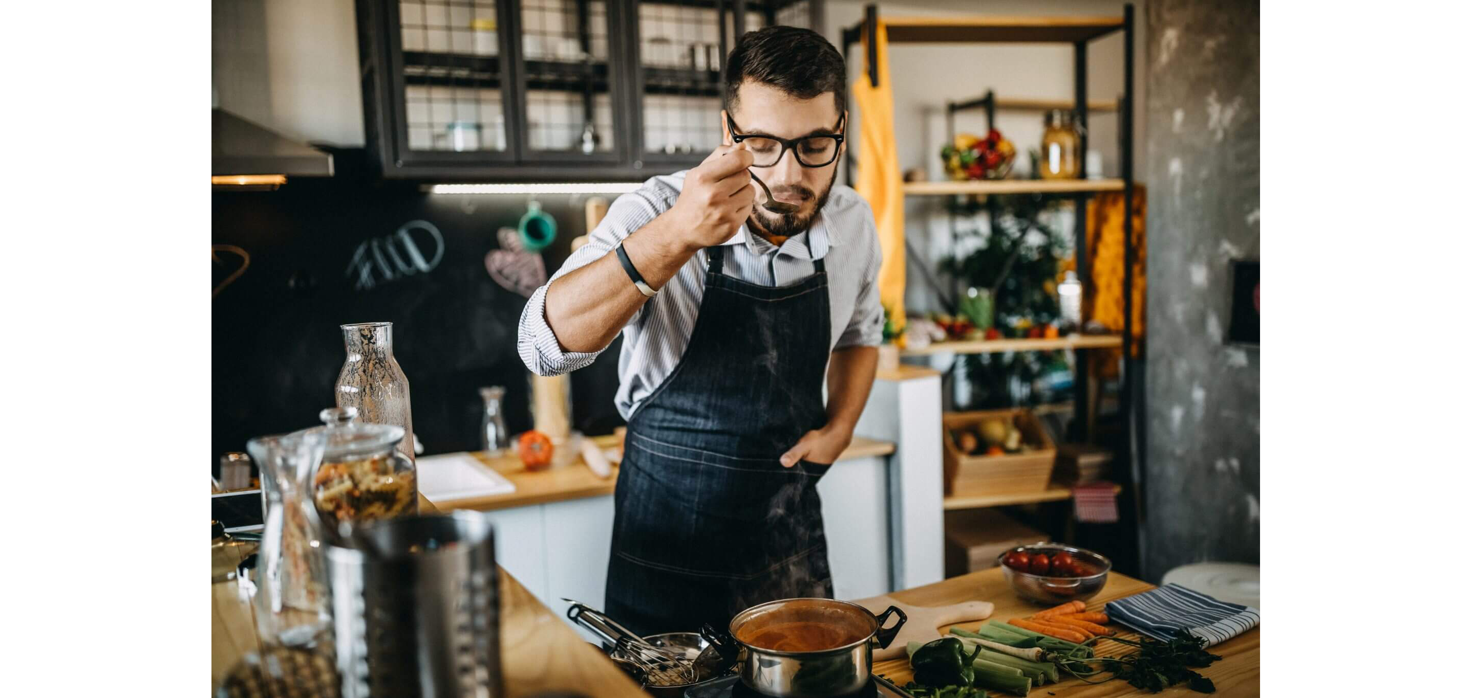 A young man in a denim apron taste-testing his homemade sauce in a pot on the stove.