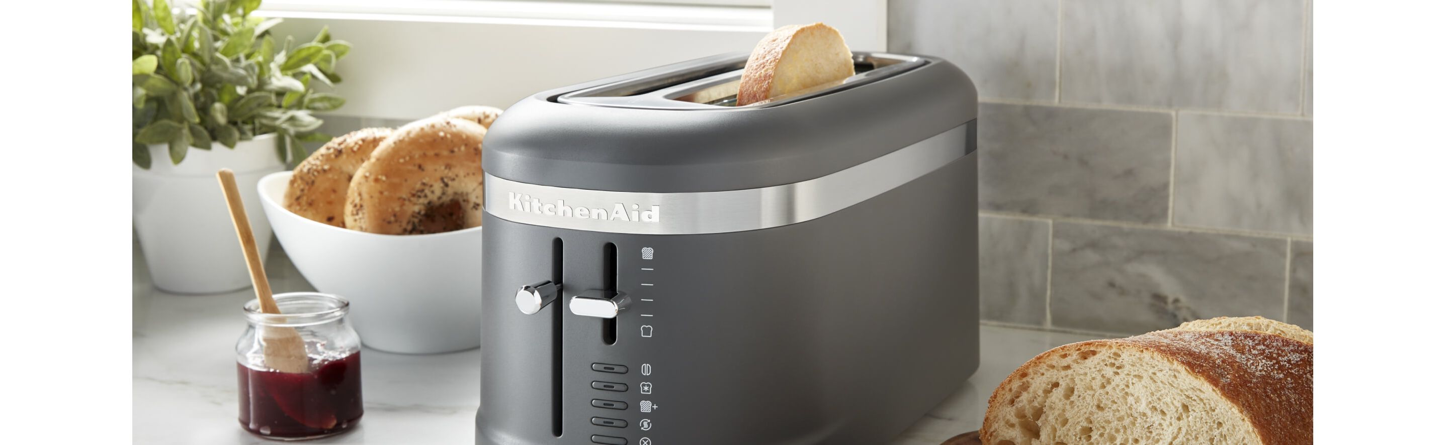 A matte black toaster on a counter with bagels, bread and jam