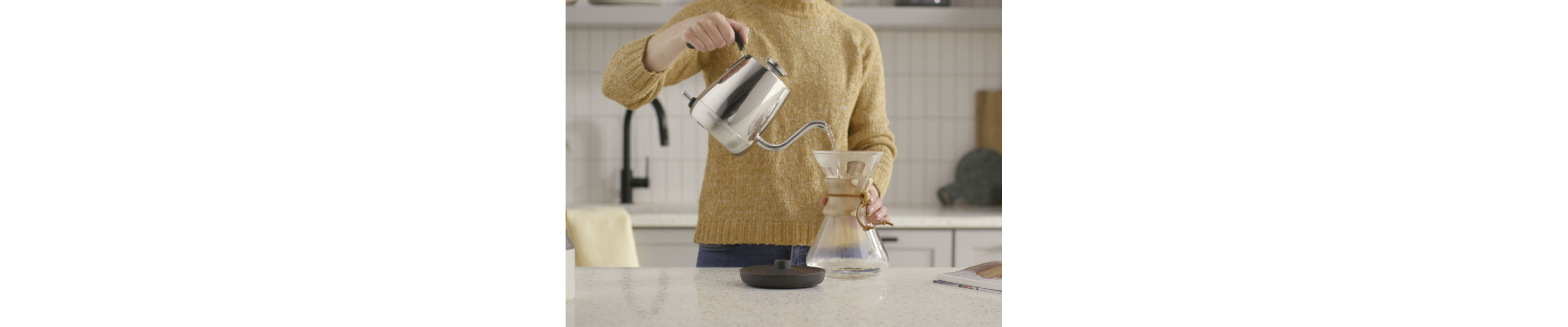A woman in a modern kitchen pouring water from a KitchenAid® electric kettle into a glass container