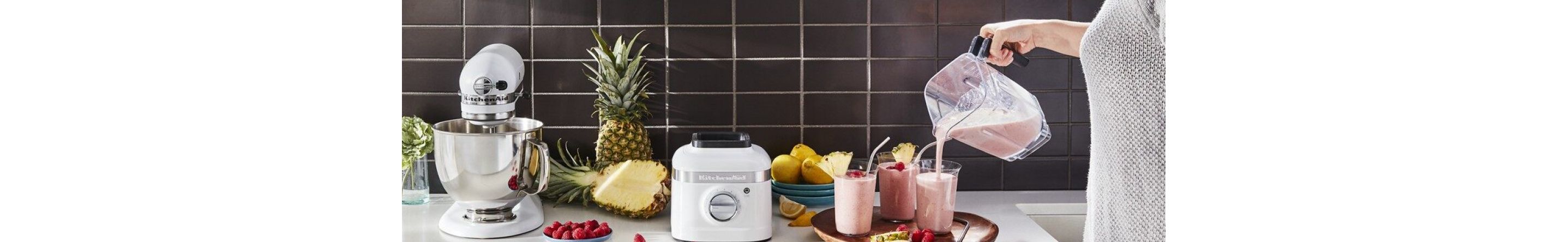 White countertop with KitchenAid® stand mixer and KitchenAid® blender surrounded by fruits - person is pouring smoothies into three glasses