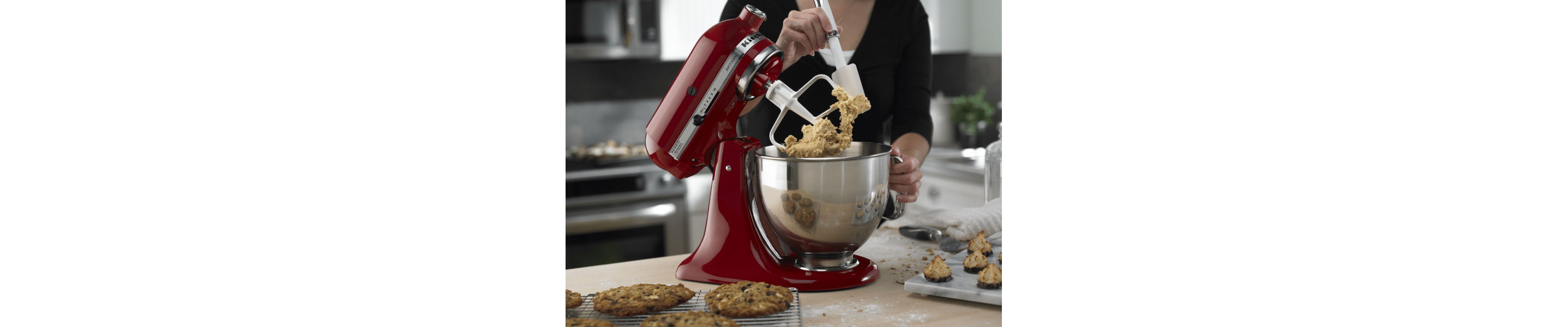 Tilt-head red KitchenAid® Stand Mixer with paddle attachment being scraped down while mixing up cookie dough with baked cookies in the foreground 